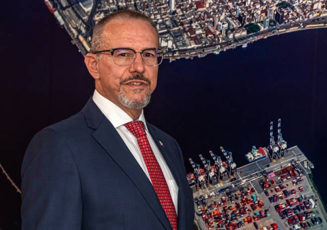 Retrato de Fabio Siccherino, Diretor-presidente da DP World, usando tenro e óculos, com painel mostrando o porto de Santos ao fundo